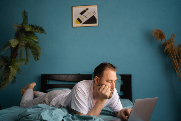Красивый белый мужчина смотрит что-то с помощью ноутбука, когда лежит на кровати в спальне. Копирование пространства - Фото, изображение