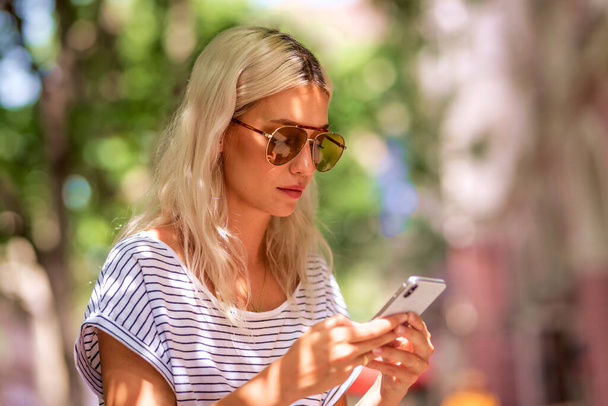Attractive blond haired woman walking on the street and text messaging. Confident female wearing striped shirt and sunglasses.  - Photo, Image