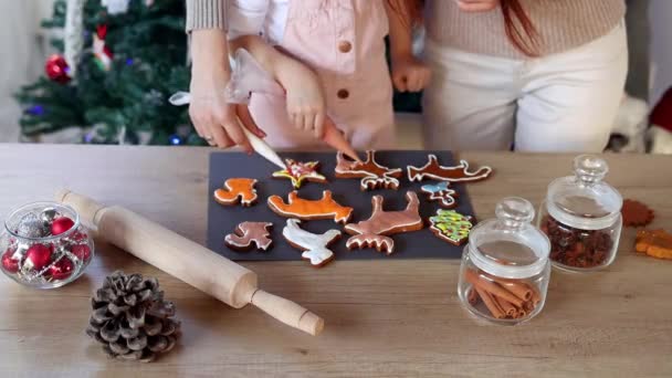 Happy little girl with her beautiful mom coloring Christmas gingerbread cookies at home. Christmas decoration in the kitchen. Fir tree with fairy lights. The concept of the New Year and Christmas - Footage, Video