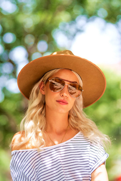 Close-up portrait of attractive blond haired girl wearing sunglasses and hat while sitting outdoor on the street.  - Fotografie, Obrázek