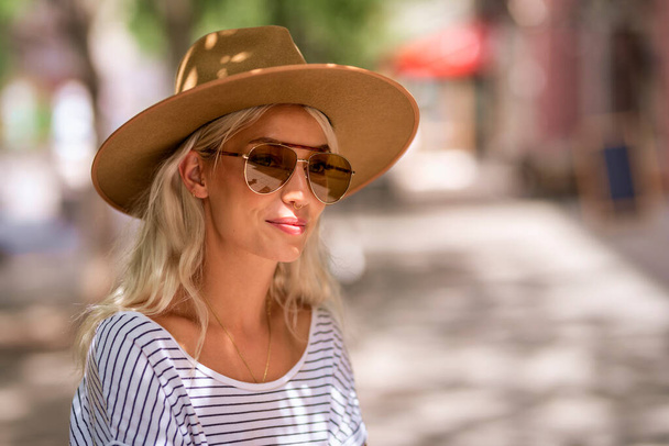 Close-up portrait of attractive blond haired girl wearing sunglasses and hat while sitting outdoor on the street.  - Φωτογραφία, εικόνα