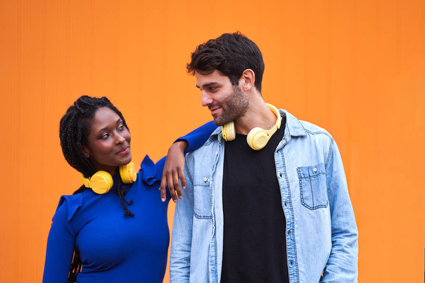 Multi-ethnic couple with headphones looking at each other while standing against a colour background. Relationship concept. - Фото, зображення