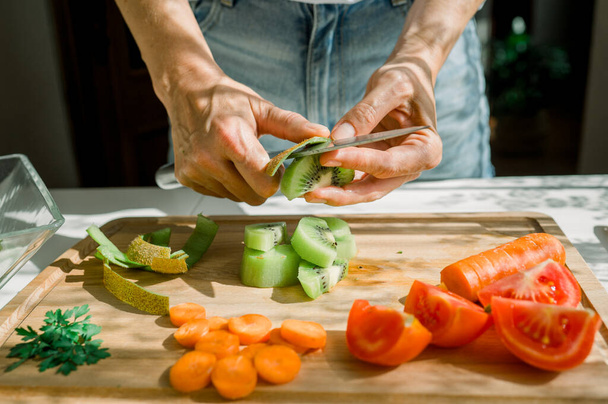 Erntehelferin schält saftige Kiwi mit Messer auf Holzschneidebrett, während sie Zutaten für gesunden Salat in der Küche zubereitet - Foto, Bild