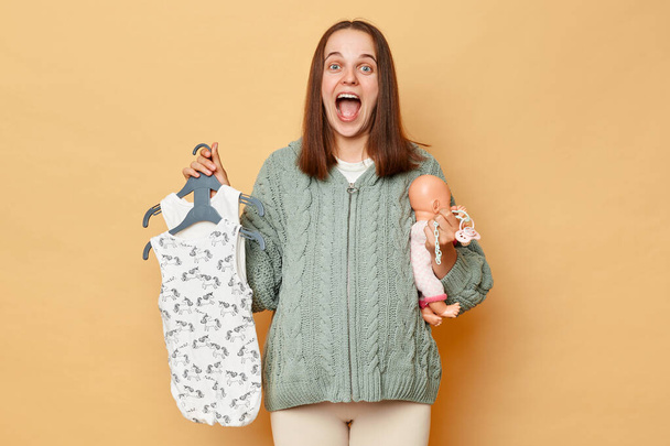 Overjoyed excited amazed young pregnant woman with baby bodysuits and doll isolated over beige background looking at camera and screaming with happy satisfied face. - Fotó, kép