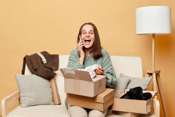happy laughing woman sitting on sofa among boxes with clothing talking on mobile phone being in good mood boasting her online shopping posing against beige wall. - Foto, afbeelding