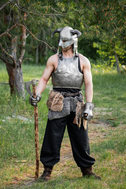 Portrait of a warrior in a horned helmet, a steel breastplate with a two-handed ax in his hands, posing against the backdrop of the forest.  - Fotó, kép