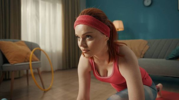 A redhaired young woman in sportswear is doing a warm up on a sports mat in the living room. A woman in a pink tracksuit and headband looks straight ahead, concentrating on the task at hand. Close up - Foto, Imagem