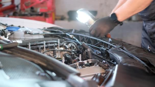 A mechanic looks under the hood of a car. A man in a gray uniform is repairing a car indoors. Close up - Materiaali, video