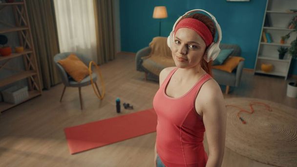 Muotokuva punatukkainen nainen valkoinen langattomat kuulokkeet olohuoneessa urheiluvälineet lattialla. Nuori nainen vaaleanpunaisessa verryttelypuvussa katsoo suoraan eteenpäin. Sulje se. - Valokuva, kuva