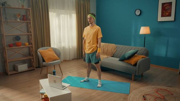 Sports at home. A man in a sportswear and with a green headband on his head stands in front of a laptop. A man watches a video training lesson. Home fitness concept - Photo, image