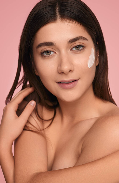 Self assured young topless female model with long dark hair and facial cream on cheek, covering breast with arms and looking at camera against pink background - Фото, зображення