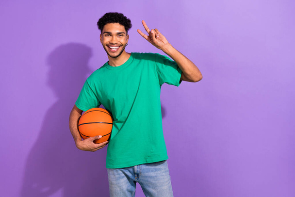 Photo of young handsome guy wear green t-shirt show v-sign hold ball professional basketball player isolated on violet color background. - Фото, изображение