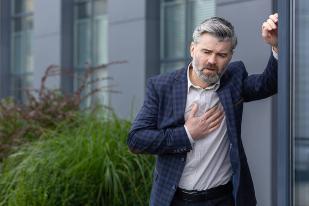 Heart attack and chest pain, mature gray-haired man outside office building has severe pain, businessman boss holding hand to chest and breathing hard from pain. - Valokuva, kuva