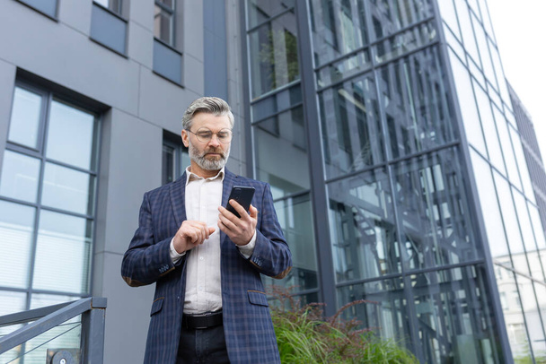 Serious thoughtful businessman boss is using an application on a smartphone, a man in a suit outside an office building is holding a phone in hands, typing a text message, and browsing, reading news. - Photo, Image
