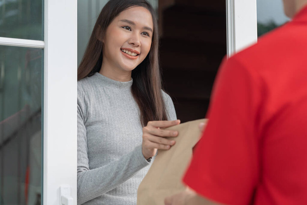 Gelukkig glimlachende Aziatische vrouw ontvangt papieren zak met voedsel van koerier voorhuis. Bezorger verzenden bezorg express. online winkelen, papieren container, afhaalservice, postbode, bezorgservice, pakketten - Foto, afbeelding