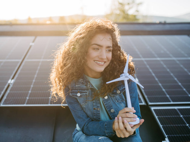 Portrait of young woman, holding model of a wind turbine on roof with solar panels. - Foto, Imagen