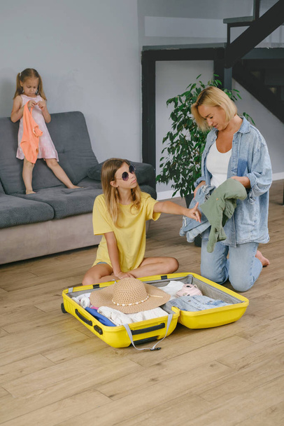 Mother with daughters packing yellow suitcase for travel. Family collecting clothes and accessories for trip in luggage. Happy and bored child getting ready for vacation. - Photo, image