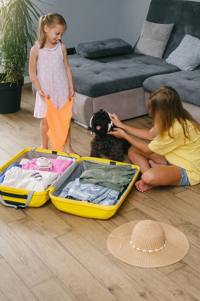 Children packing suitcase for travel and play with a dog. Sisters having fun with pet while getting ready for a trip. Dog in sunglasses and sunhat. going on vacation - Фото, зображення