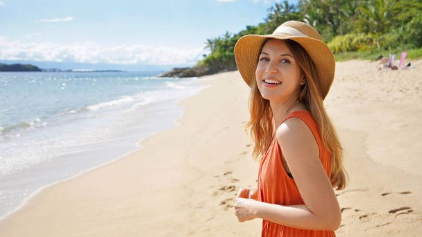 Foto panorámica de una chica sonriente con sombrero de paja y vestido girando sin mirar a la cámara en la hermosa playa tropical soleada vacía. Anuncio de banner y espacio de copia. - Foto, Imagen