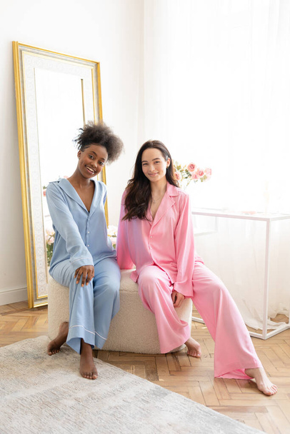 Coppia di donne africane e caucasiche sorridenti in pigiama in posa seduta in poltrona in camera da letto sullo sfondo degli interni di casa. - Foto, immagini