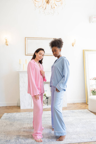 Couple of african and caucasian women in pajamas posing standing on carpet in bedroom against background of home interior. - Photo, image