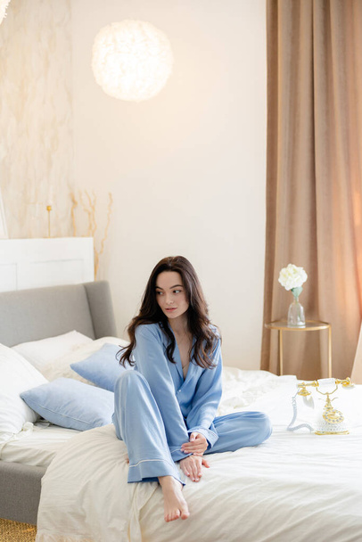 Jeune femme brune coûteuse en pyjama bleu assis sur le lit sur fond de linge de lit et l'intérieur de la maison. - Photo, image
