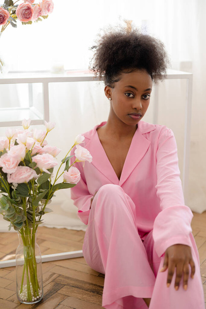 Młoda afrykańska zamyślona kobieta w różowej piżamie siedząca na podłodze w pobliżu bukietu kwiatów na tle okna. - Zdjęcie, obraz