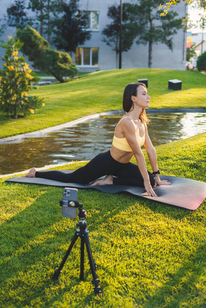 Vue latérale de la jeune femme en tenue de sport assise sur un tapis et utilisant un smartphone tout en enregistrant une vidéo pour une session de yoga - Photo, image