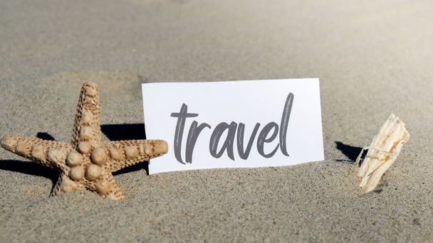 ТРАВЕЛ текст на бумажной поздравительной открытке на фоне летнего декора раковины морской звезды. Побережье песчаного пляжа. Открытка с праздничной концепцией. Концепция туристического бизнеса - Фото, изображение
