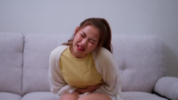 Beteg ázsiai nő szenved akut hasi fájdalom a has miatt menstruációs időszak, PMS. Ül a kanapén, gyomorfájás ételmérgezés, hasi fájdalom, emésztési probléma, gyomorhurut - Felvétel, videó