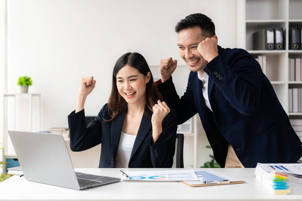 Dos jóvenes empresarios asiáticos exitosos levantando las manos con alegría con computadoras portátiles. Celebren juntos victorias y éxitos. Aplausos en la oficina moderna - Foto, Imagen