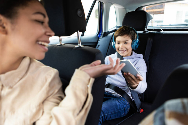 Hymyilevä musta nainen antaa älypuhelimen pojalleen pelata autossa. Perheen, kuljetuksen, matkan, matkustamisen ja ihmisten käsite - Valokuva, kuva