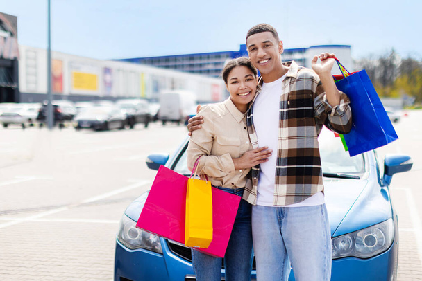 African couple with shopping bags smiling and standing near blue car in store parking. Love, casual lifestyle or shopaholic concept. With copy space. High quality photo - Valokuva, kuva