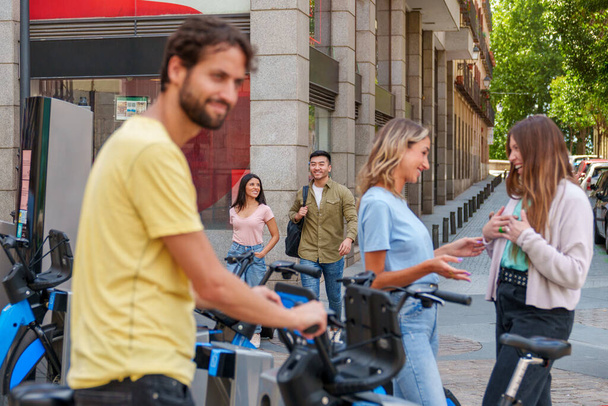 Μια ποικιλόμορφη ομάδα νέων ανθρώπων, που εκπροσωπούν διάφορες εθνότητες, συγκεντρώνεται σε ένα χώρο στάθμευσης της πόλης για μια συναρπαστική ημέρα ενοικίασης ηλεκτρικών ποδηλάτων κάτω από τον ζεστό ήλιο. - Φωτογραφία, εικόνα