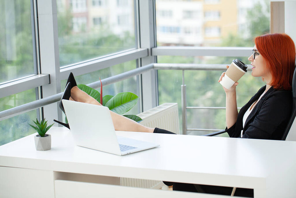 Mulher com cabelo vermelho está bebendo café e falando ao telefone no local de trabalho no escritório. - Foto, Imagem