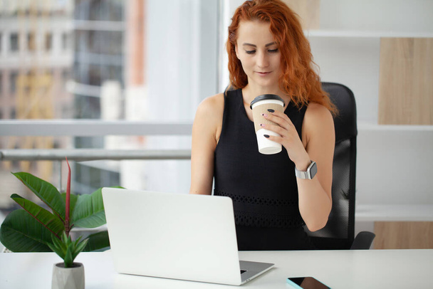 Женщина с рыжими волосами пьет кофе и разговаривает по телефону на рабочем месте в офисе. - Фото, изображение