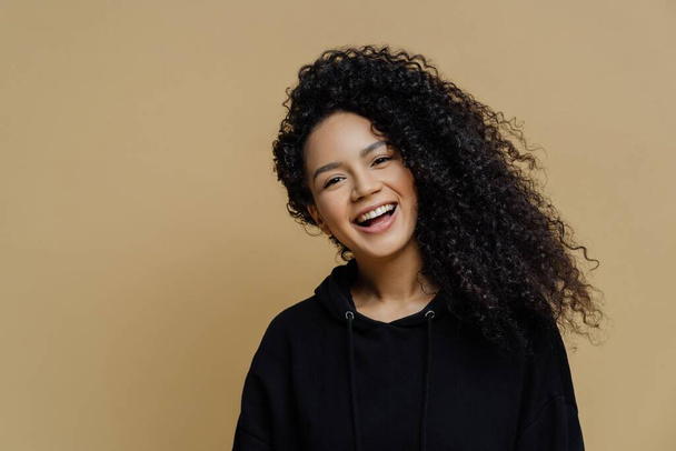 Positive African American woman smiles, tilts head happily, wearing black sweatshirt, isolated on beige background, radiating joy. Happiness personified. - Foto, immagini