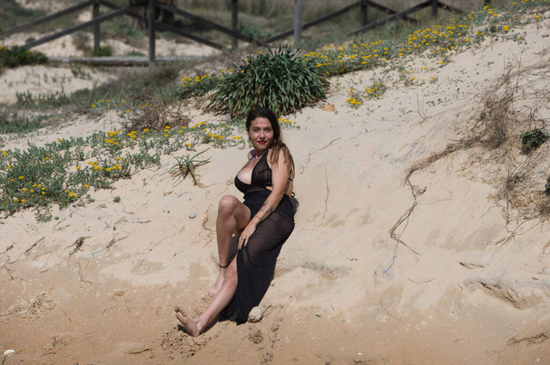 Красивая молодая женщина с длинными каштановыми волосами в купальнике и черным шелковым саронгом лежит на дюнах пляжа, позируя чувственно для фотографий. Женщина очень красивая.. - Фото, изображение