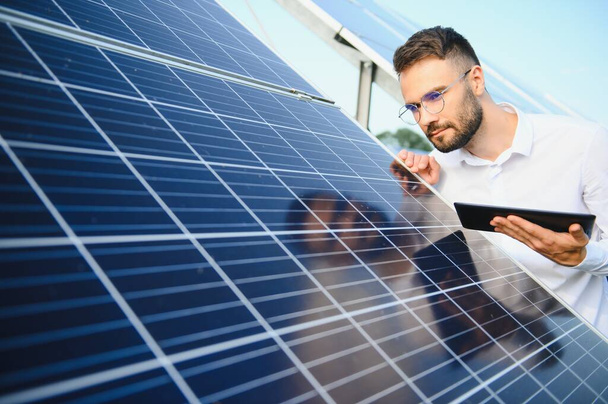 Bearded male engineer using tablet to control photovoltaic panels remotely during work on solar farm on sunny day. - 写真・画像