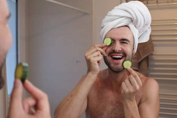 retrato de hombre joven con toalla en la cabeza y pepinos en los ojos delante del espejo en el baño - Foto, Imagen