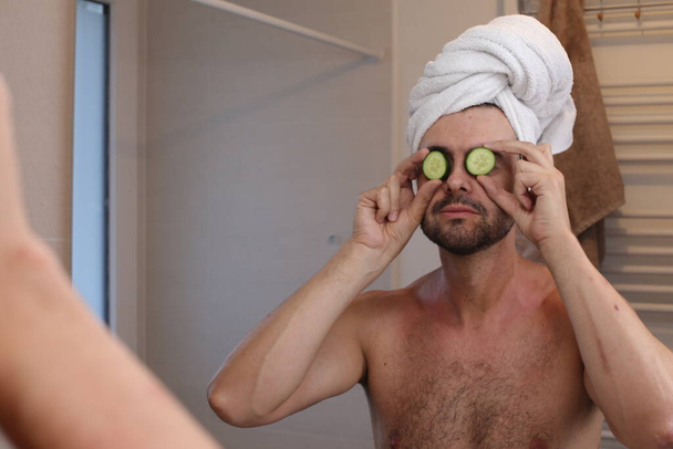 retrato de hombre joven con toalla en la cabeza y pepinos en los ojos delante del espejo en el baño - Foto, Imagen