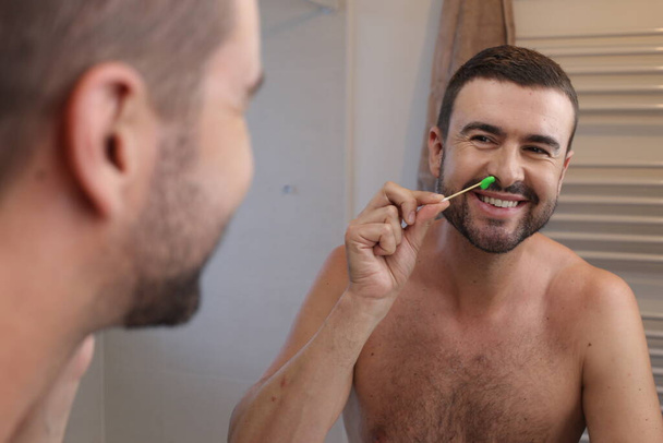 портрет молодого человека, натирающего волосы на носу перед зеркалом в ванной комнате - Фото, изображение