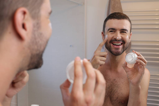 πορτρέτο του νεαρού άνδρα με βάζο ενυδατική κρέμα μπροστά από τον καθρέφτη στο μπάνιο - Φωτογραφία, εικόνα