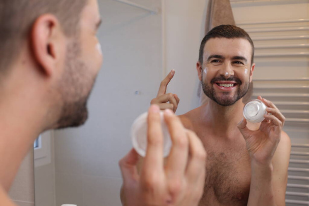portrait of young man with jar of moisturizing cream in front of mirror in bathroom - Foto, Imagen