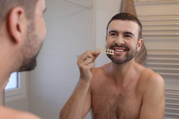 portrait of young man with plastic teeth tone samples in front of mirror in bathroom - Photo, Image