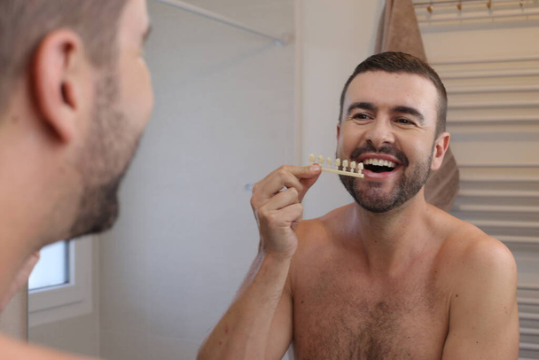 portrait of young man with plastic teeth tone samples in front of mirror in bathroom - Photo, Image