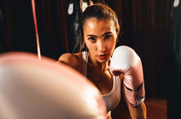 Femme asiatique Muay boxer thaïlandais poing poing devant la caméra en position de combat prêt à poser à la salle de gym avec équipement de boxe en arrière-plan. Regard de détermination concentré et préparation au défi. Impulsion - Photo, image