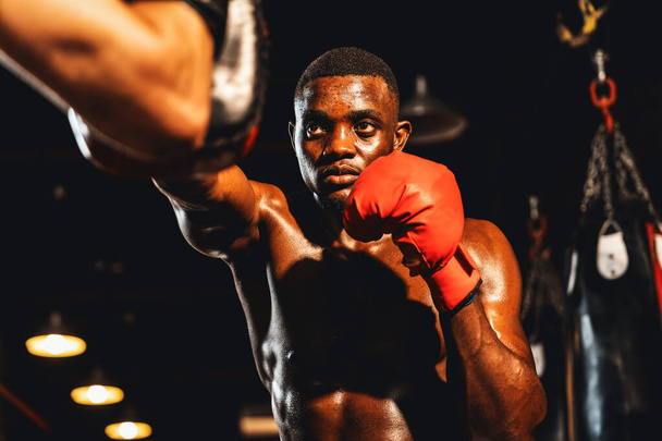 アフリカ系アメリカ人のブラックボクサーは、ジムでボクシングバッグトレーニング機器としてパンチングミットを身に着けてトレーナーやコーチでパンチ。プロボクシングの試合のための強さとスタミナトレーニング。インペトゥス - 写真・画像