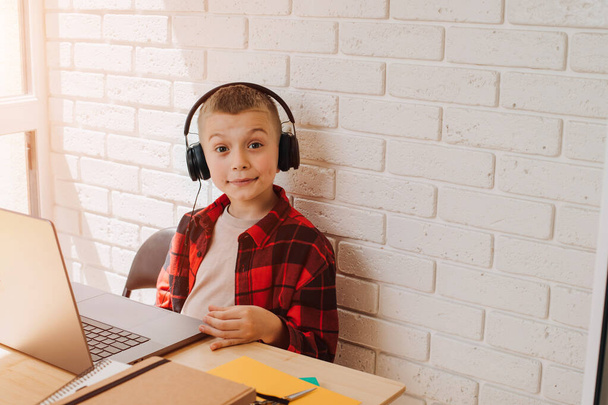 Školní kluk v černých sluchátkách sedí u stolu s laptopem. Děti a přístroje. Pohled zepředu - Fotografie, Obrázek