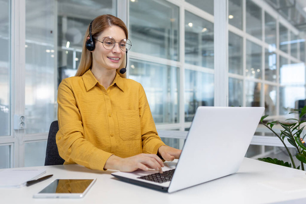 Smiling businesswoman working inside office with laptop and headset for video call, woman sitting at workplace happy working with clients - Фото, зображення